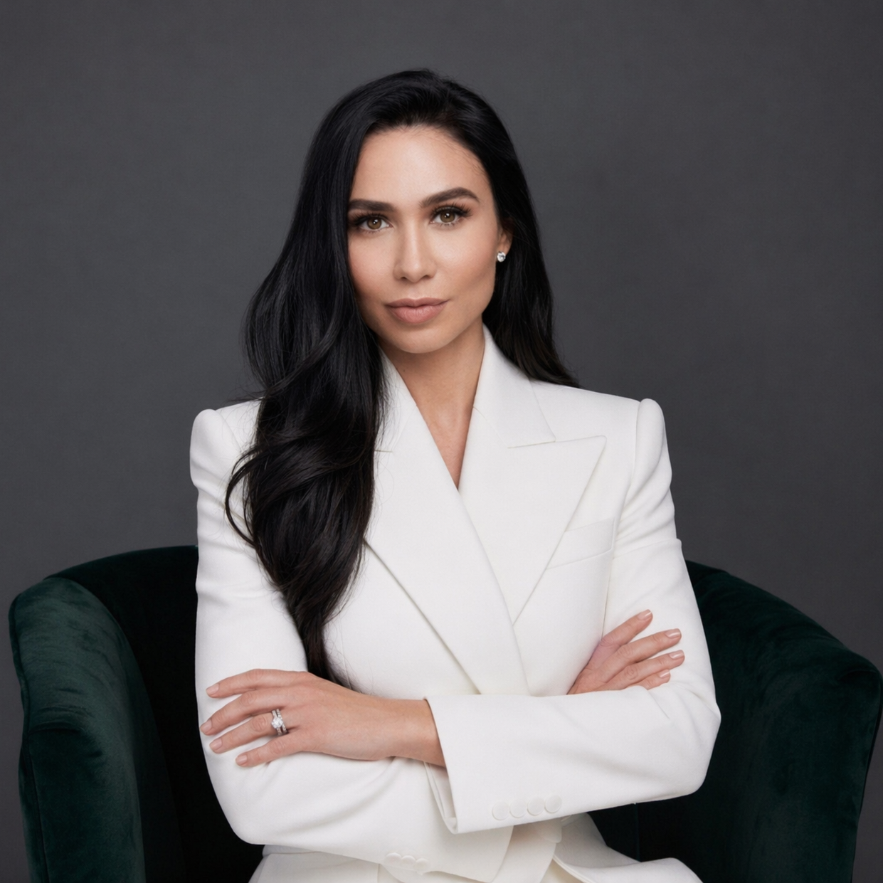 Woman founder of Beleza by Z professional tallow skincare posing in a white suit while sitting on a dark green velvet chair against a gray background